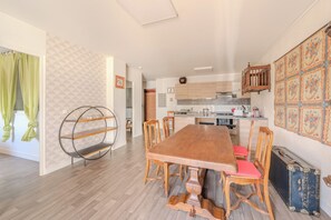 A dining area with a wooden table and chairs, a kitchen in the background, and a patterned wall. - La Grande Terrasse - BBQ - Proche Golf (Gargenville)