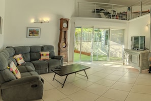 A living room with a sectional sofa, a coffee table, a clock, a TV, and a balcony with a railing. - Kaz vert - Piscine - Wifi (Saint-Pierre)