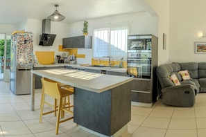A modern kitchen with a central island, bar stools, a built-in oven, and a grey sofa in the living area. - Kaz vert - Piscine - Wifi (Saint-Pierre)
