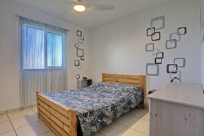 A bedroom with a wooden bed, a bedside table, a nightstand, a window with curtains, and a wall with geometric patterns. - Kaz vert - Piscine - Vue mer (Saint-Pierre)