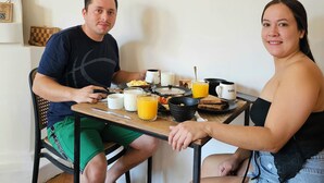 Zwei Personen sitzen an einem Tisch mit Frühstück, einem Laptop und einem Smartphone.