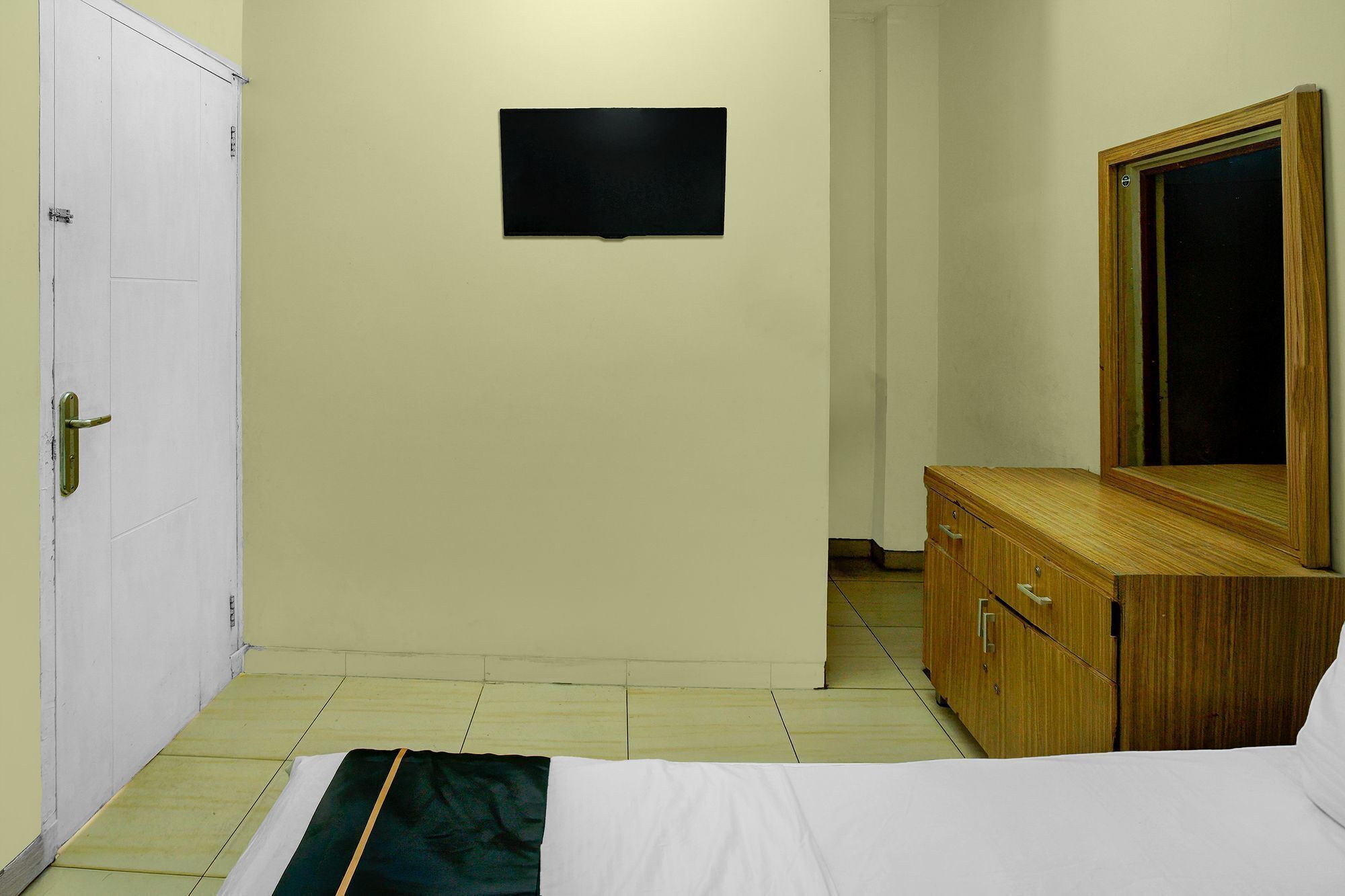 A hotel room with a bed, a wooden dresser, a television mounted on the wall, and a door.