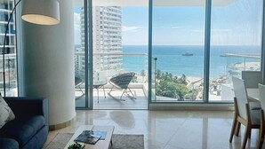 En rummelig stue med en stor skydedør, der fører ud til en balkon med havudsigt og udsigt over bybilledet.