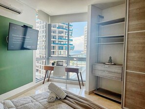 A modern hotel room with a large window, a desk, a chair, a bed, and a wardrobe. - 2BR: Modern Ocean View Apartment | Grand Marina (Santa Marta)