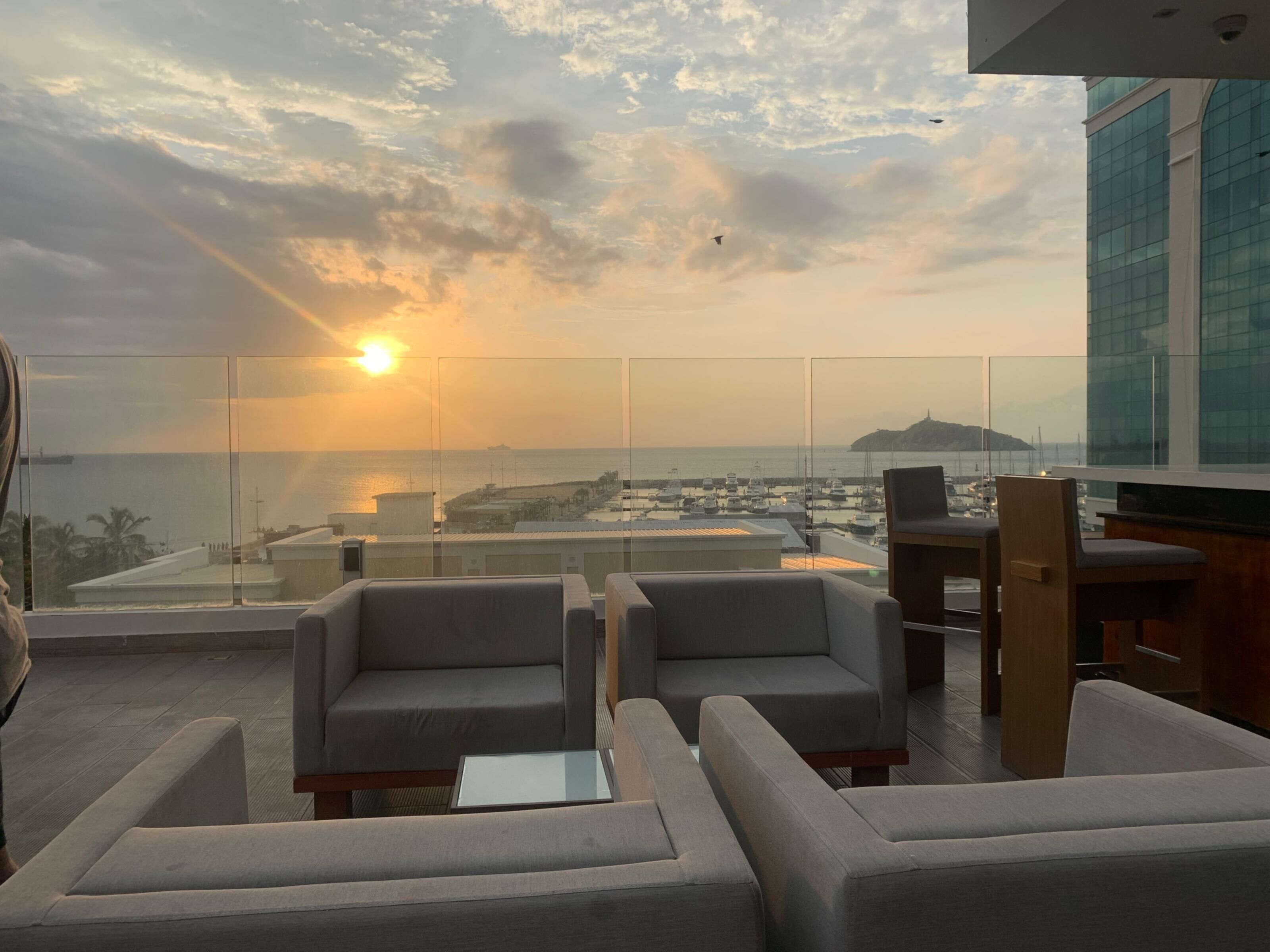 A rooftop lounge with seating and a view of the sea and cityscape during sunset.
