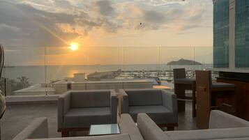A rooftop lounge with seating and a view of the sea and cityscape during sunset.