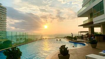 A rooftop pool with clear views of the sunset and ocean, surrounded by lounge chairs and potted plants.