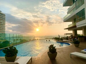 Ein Pool auf dem Dach mit freiem Blick auf Sonnenuntergang und Meer, umgeben von Liegestühlen und Topfpflanzen.