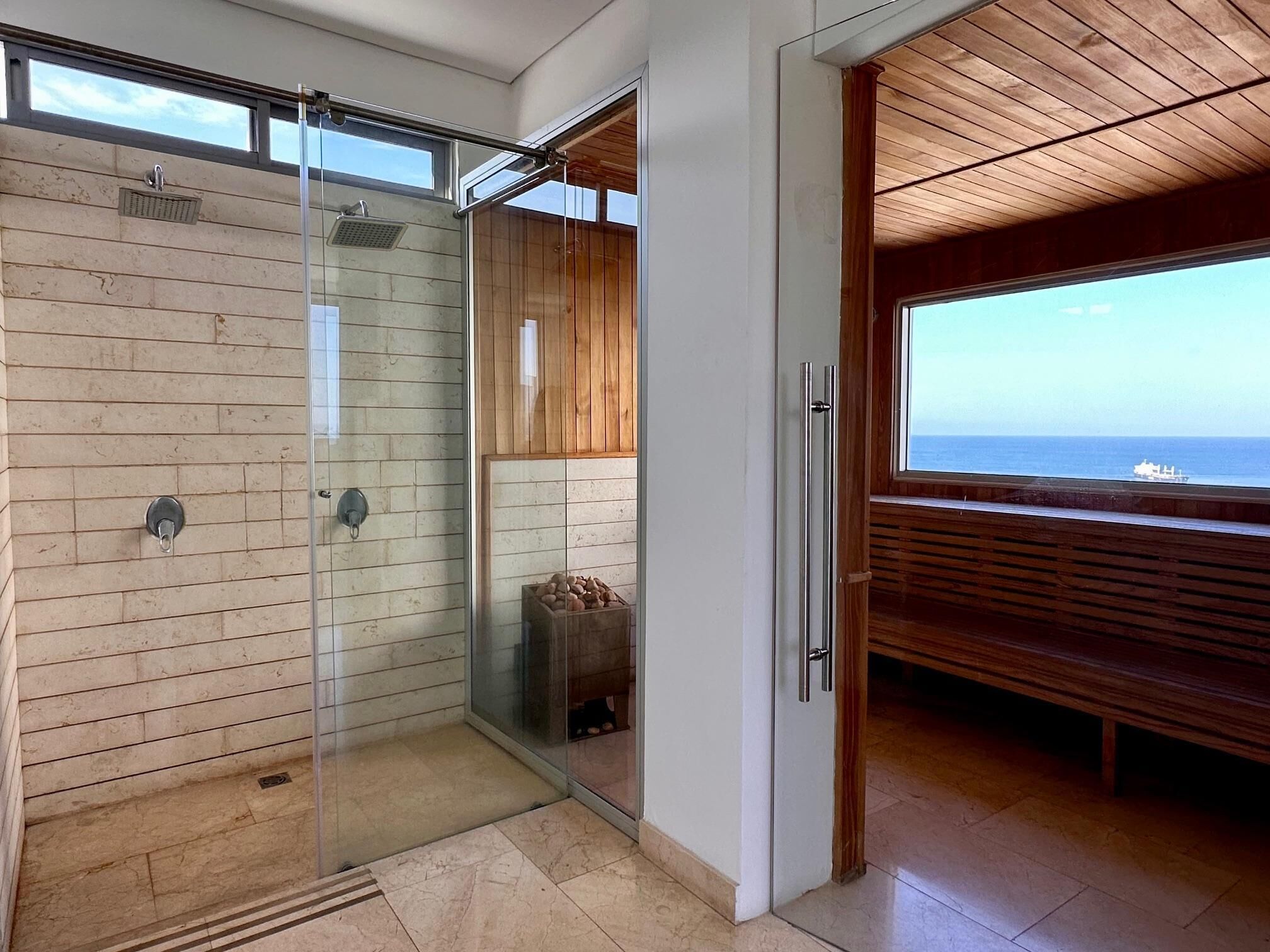 A modern bathroom with a glass shower, wooden paneling, and a view of the sea.