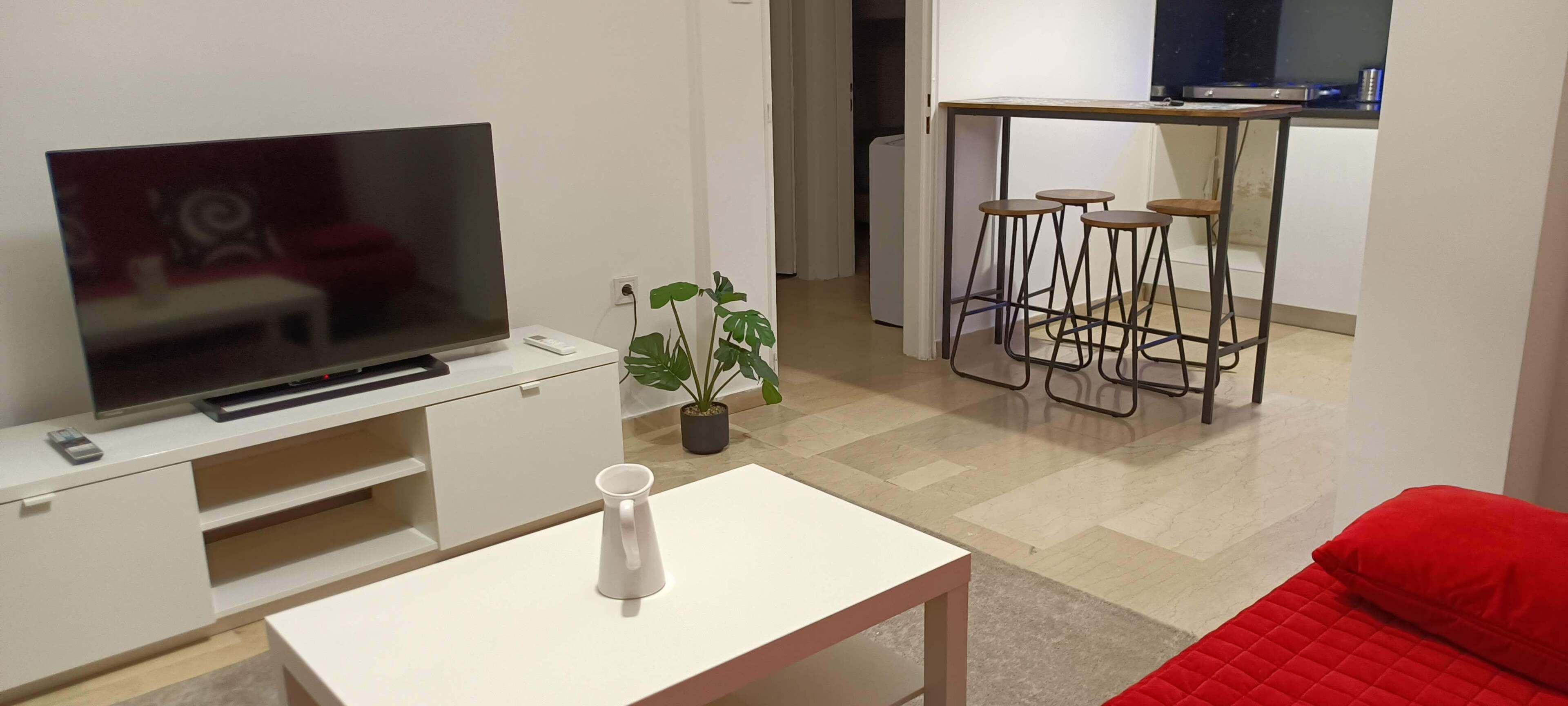 A living room with a flat-screen TV, a white coffee table, a red couch, a small plant, and a kitchen area with stools.