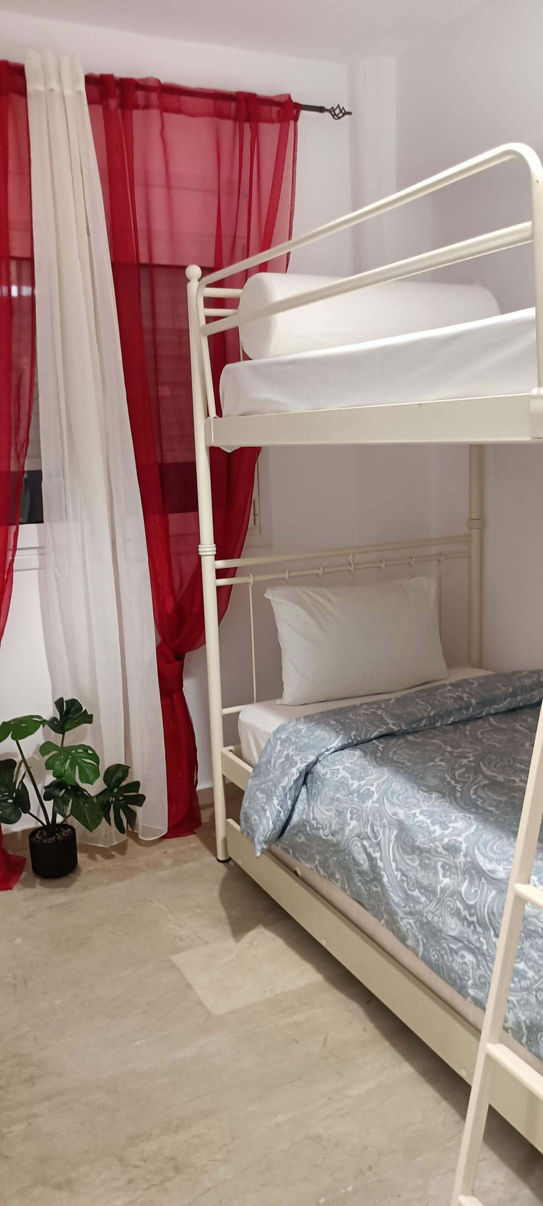 A bunk bed with white bedding, a potted plant, and red and white curtains.