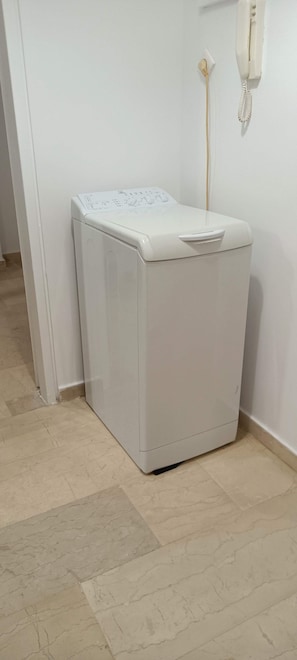 A white washing machine in a corner of a room with a tiled floor and a wall-mounted telephone. - Marousi Accomodation 2 bedrooms (Marousi)