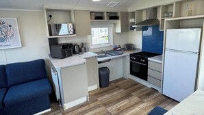 Una kitchenette compacta con estufa, lavamanos y refrigerador, un sofá azul y un ventanal con vistas a un edificio.