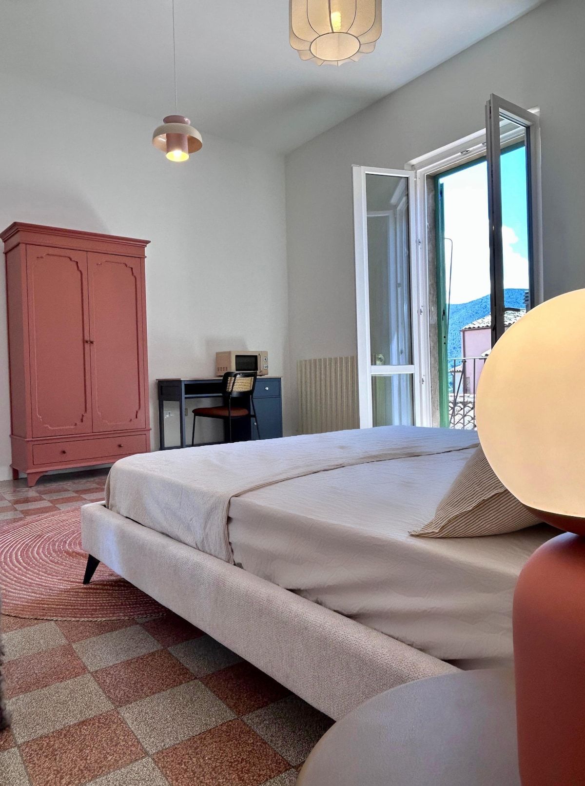 A bedroom with a bed, a desk, a chair, a wardrobe, and a view through an open door.