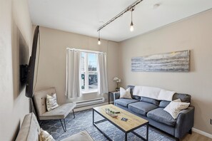 Ein Wohnzimmer mit einer Couch, einem Couchtisch, einem an der Wand montierten Fernseher, einem Fenster mit Vorhängen und einem Gemälde an der Wand.