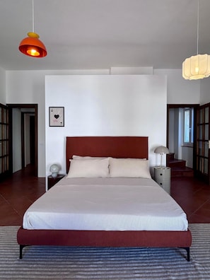 Ein Schlafzimmer mit einem großen Bett, einem Nachttisch, einer Nachttischlampe, einem gerahmten Bild an der Wand und einer Tür zu einem anderen Raum.