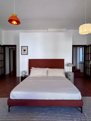 Une chambre à coucher avec un grand lit, une table de chevet, une commode, un tableau accroché au mur et deux lampes suspendues.