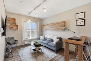 A modern living room with a grey sofa, a wooden coffee table, a black chair, a standing lamp, and a wall-mounted shelf. - Stateview 203 - 1BR + Free Co-Working Space (Appleton)