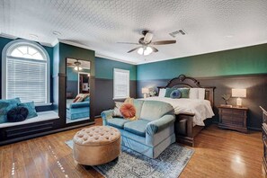 Ein Schlafzimmer mit einem Bett, einem blauen Sofa, einem runden Hocker, einem Deckenventilator und einem Fenster mit Jalousien.