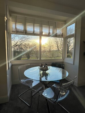 A room with a round glass table, metal chairs, a bowl with decorative items, and a view of trees through large windows. - Salvation View 2 Bed Apartment (Benfleet)