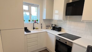 A compact kitchen with white cabinets, a black stove, and a window with plants. - Salvation View 2 Bed Apartment (Benfleet)