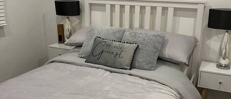 A neatly made bed with a pillow that says 'BE OUR Guest,' two bedside lamps, and a white nightstand.
