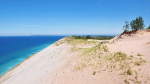 A coastal landscape with a clear sky, sandy terrain, and sparse vegetation.