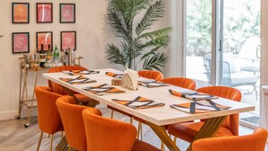 A dining area with a long table set for six, surrounded by orange chairs, a large potted plant, and framed artwork on the wall. - Mid-Century 3BR Near Banner & ASU | Extended Stay (Tempe)