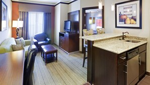 Une chambre d’hôtel avec un bureau en bois, un canapé, une chaise, une télévision et un coin cuisine équipé d’un évier, ainsi qu’un tableau encadré accroché au mur.