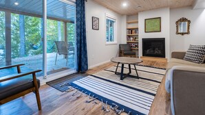 A living room with a fireplace, a sofa, a table, and a view of the outdoors. - Diamond Lakehouse | SUP, Canoe, BBQ, Aurora Magic (Highland Grove)
