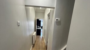 A narrow hallway with wooden flooring, white walls, and a door leading to a kitchen area. - 5BR Home w/ Parking for Groups & Contractors (Portsmouth)