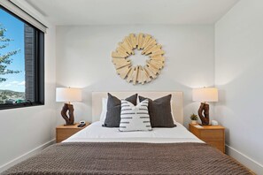 A modern bedroom with a large bed, two bedside tables with lamps, a wall-mounted abstract art piece, and a view of the outdoors through a window. - Ski-In 5BR Canyons Hideaway | Hot Tub, Ski Storage | Steps to Gondola (Park City)