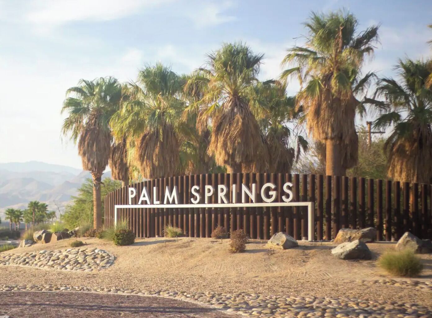 A sign for Palm Springs with a wooden fence and palm trees in the background.