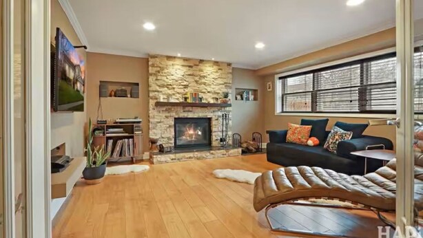 A living room with a stone fireplace, a black sofa with colorful cushions, a leather armchair, and a large window with blinds.