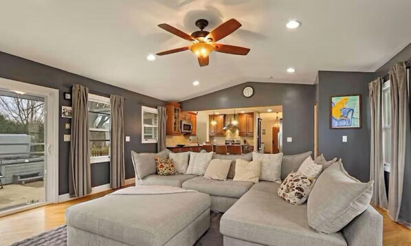 Un salon moderne avec un canapé d’angle, un ventilateur de plafond et une cuisine en arrière-plan.