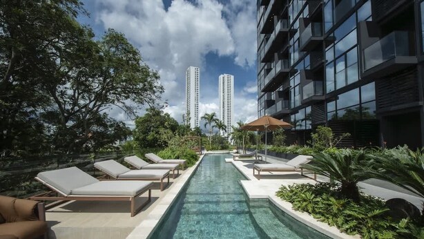 Una piscina con sillas de descanso, rodeada de vegetación y edificios altos al fondo.