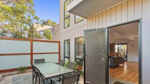 A modern outdoor patio with a glass table and chairs, a sliding glass door leading to an interior living space, and a wooden fence. - Spacious Townhouse Near Shops and Beach (Cowes)