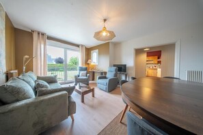 Ein großzügiges Wohnzimmer mit einem großen Esstisch aus Holz, zwei Sesseln, einer Couch und einem Fernseher. Ein Balkon bietet Blick auf Grünflächen.