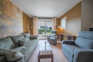 Ein Wohnzimmer mit Sofa, Sessel und Couchtisch, einer Lampe und Blick durch eine Glastür.