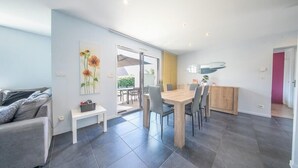 Ein modernes Wohnzimmer mit einer Couch, einem Esstisch mit Stühlen, einer Küchenzeile und einer Glastür, die auf eine Terrasse im Freien führt.