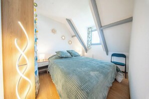 Ein Schlafzimmer mit schrägen Wänden, einem Bett mit grüner Bettdecke, einem Sessel und einer Wandleuchte.