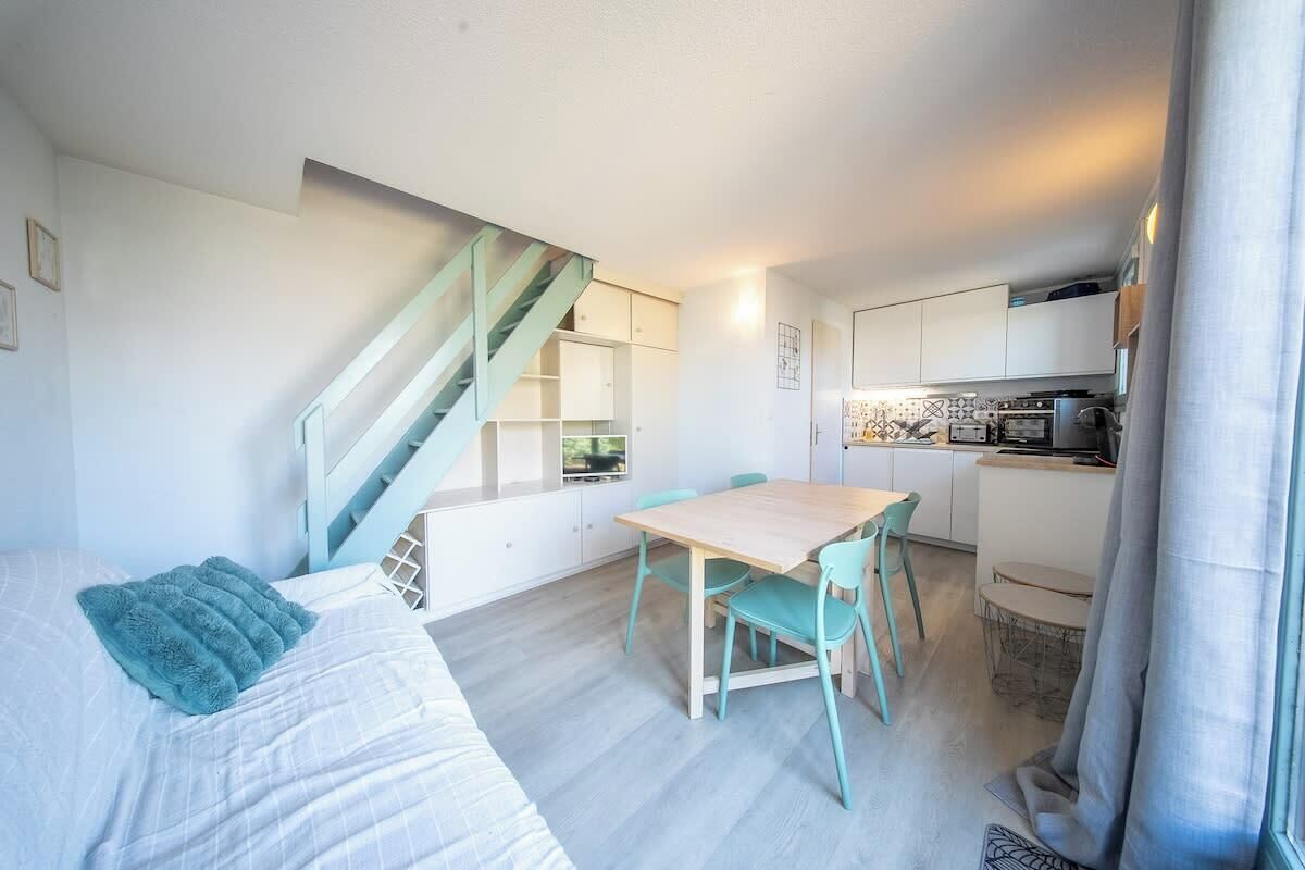 Une pièce de taille modeste comprenant un lit, une table à manger avec des chaises, un coin cuisine et un escalier.