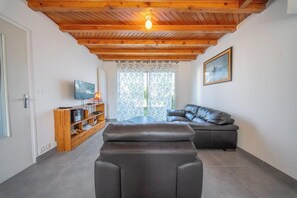 Ein modernes Wohnzimmer mit Holzboden, einem schwarzen Ledersofa, einem Holz-TV-Schrank und einem großen Fenster mit gemusterten Vorhängen.