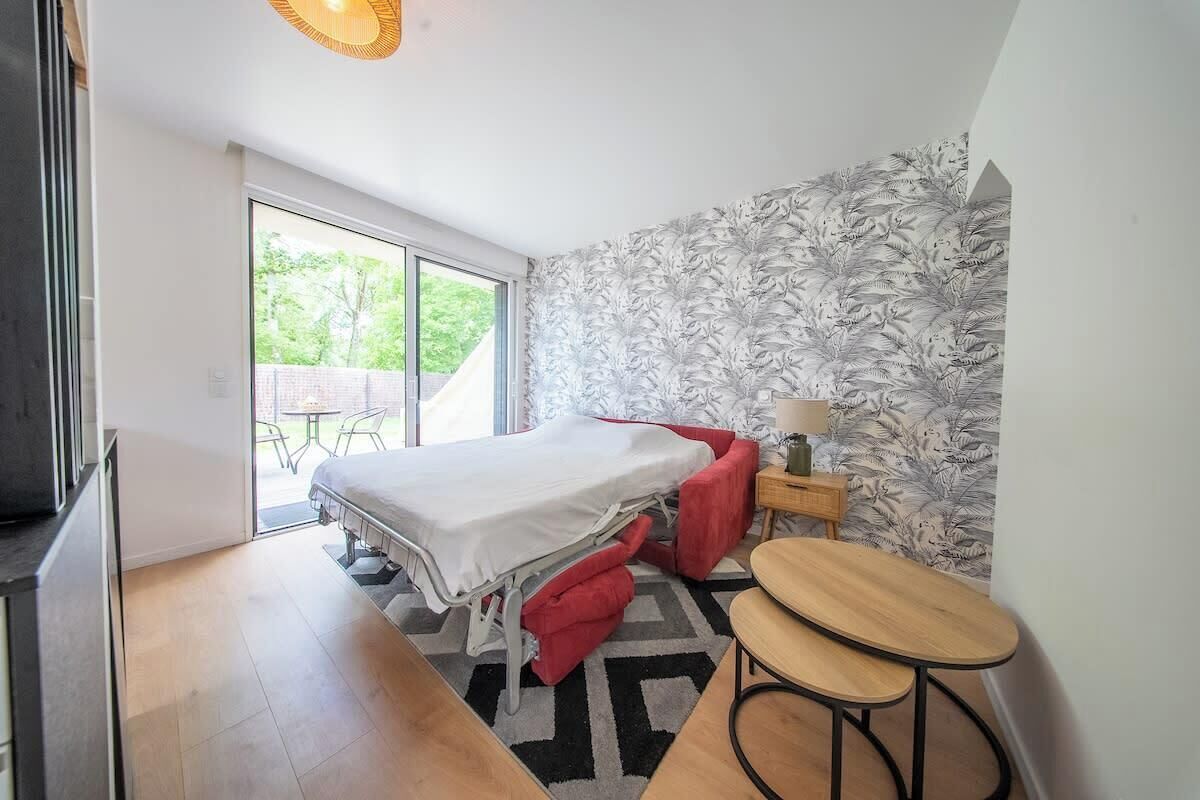 Une chambre à coucher comprenant un lit, une petite table, une chaise, une table de chevet et une porte-fenêtre coulissante donnant sur un espace extérieur.