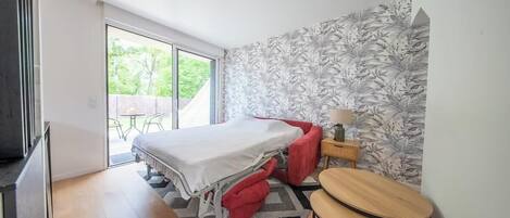 Une chambre à coucher comprenant un lit, une petite table, une chaise, une table de chevet et une porte-fenêtre coulissante donnant sur un espace extérieur.