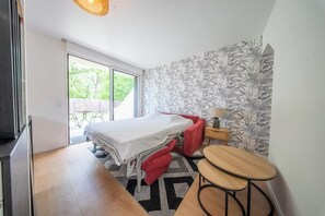 Ein Schlafzimmer mit einem Bett, einem kleinen Tisch, einem Stuhl, einem Nachttisch und einer Glastür, die zu einem Außenbereich führt.