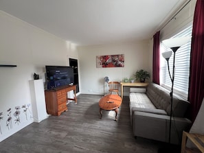 A living room with a sofa, a wooden coffee table, a TV on a stand, a desk with a chair, a wall-mounted painting, and a window with curtains.