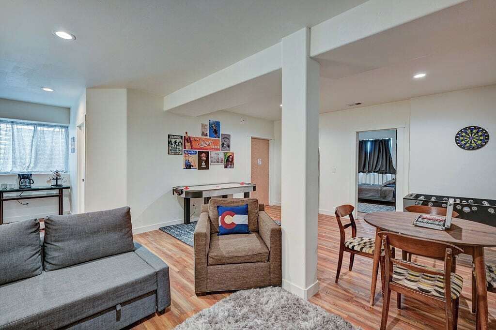 Una sala de estar con sofá, sillón y una mesa con sillas. Se observa un dardo en la pared y una mesa de futbolín al fondo.