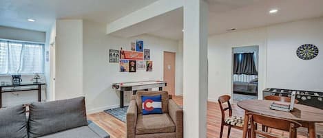 Una sala de estar con sofá, sillón y una mesa con sillas. Se observa un dardo en la pared y una mesa de futbolín al fondo.