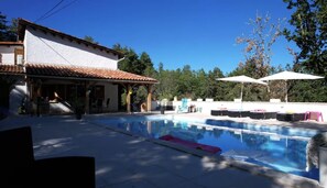 Ein Schwimmbecken mit Liegestühlen und Sonnenschirmen, ein Gebäude mit Ziegeldach und Bäume im Hintergrund.